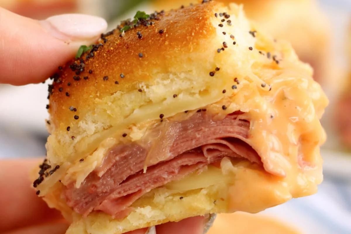 A hand holding a slider with ham and melted cheese on a poppy seed-topped bun.
