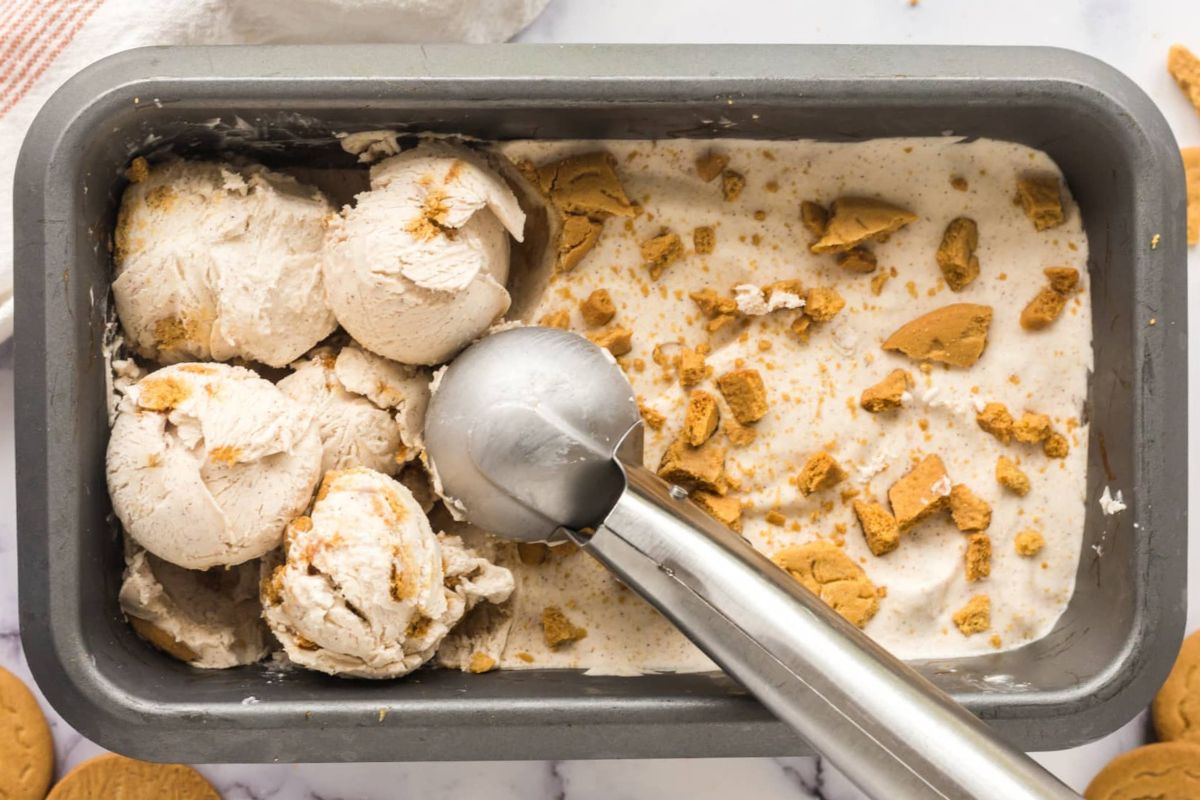 A metal scoop serving cookie-flavored ice cream recipes from a container with crumbled cookies on top.