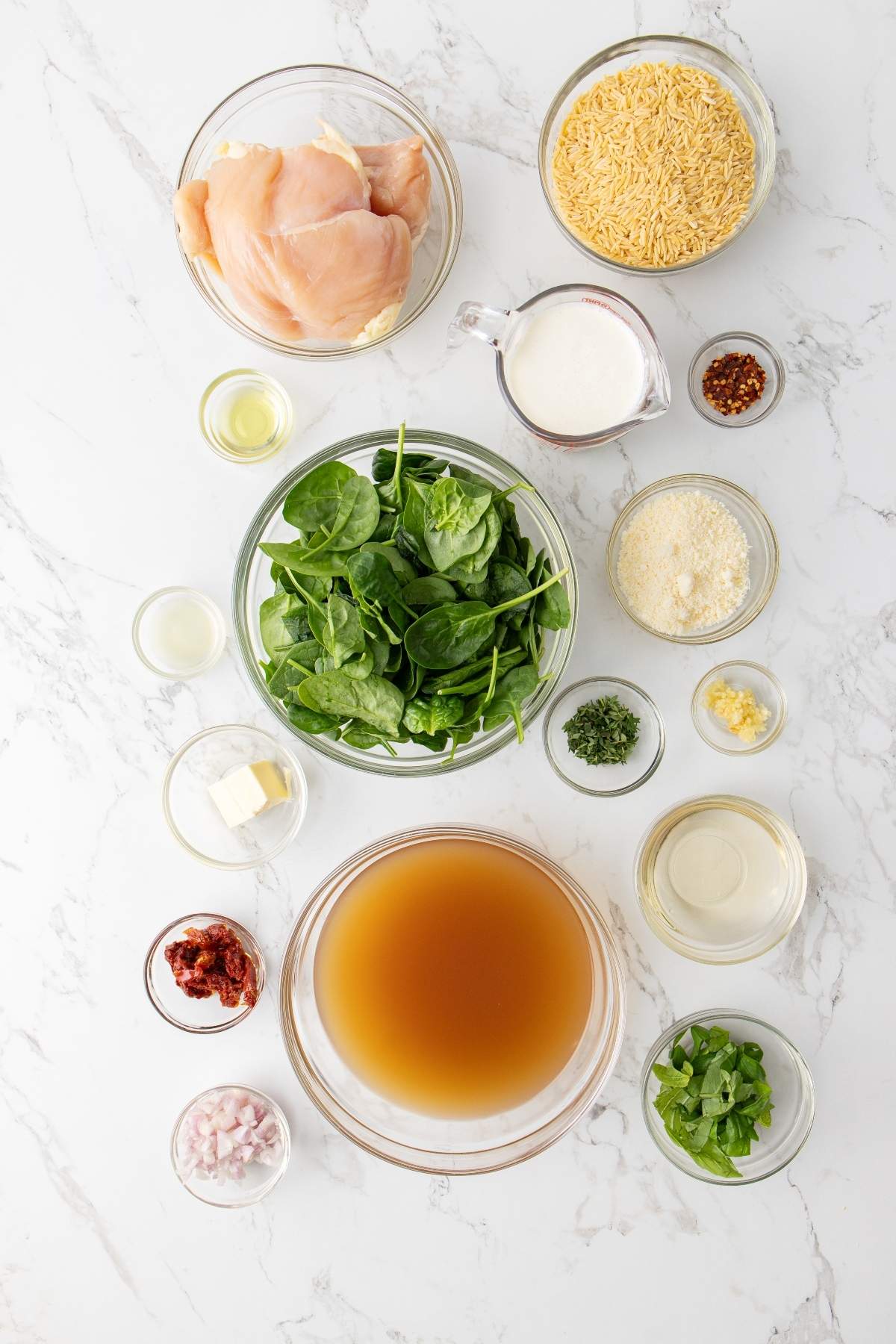 Various ingredients arranged on a marble surface, including raw chicken, fresh spinach, chicken broth, uncooked orzo pasta, milk, grated cheese, garlic, red pepper flakes, and other seasonings for an Italian chicken orzo dish.