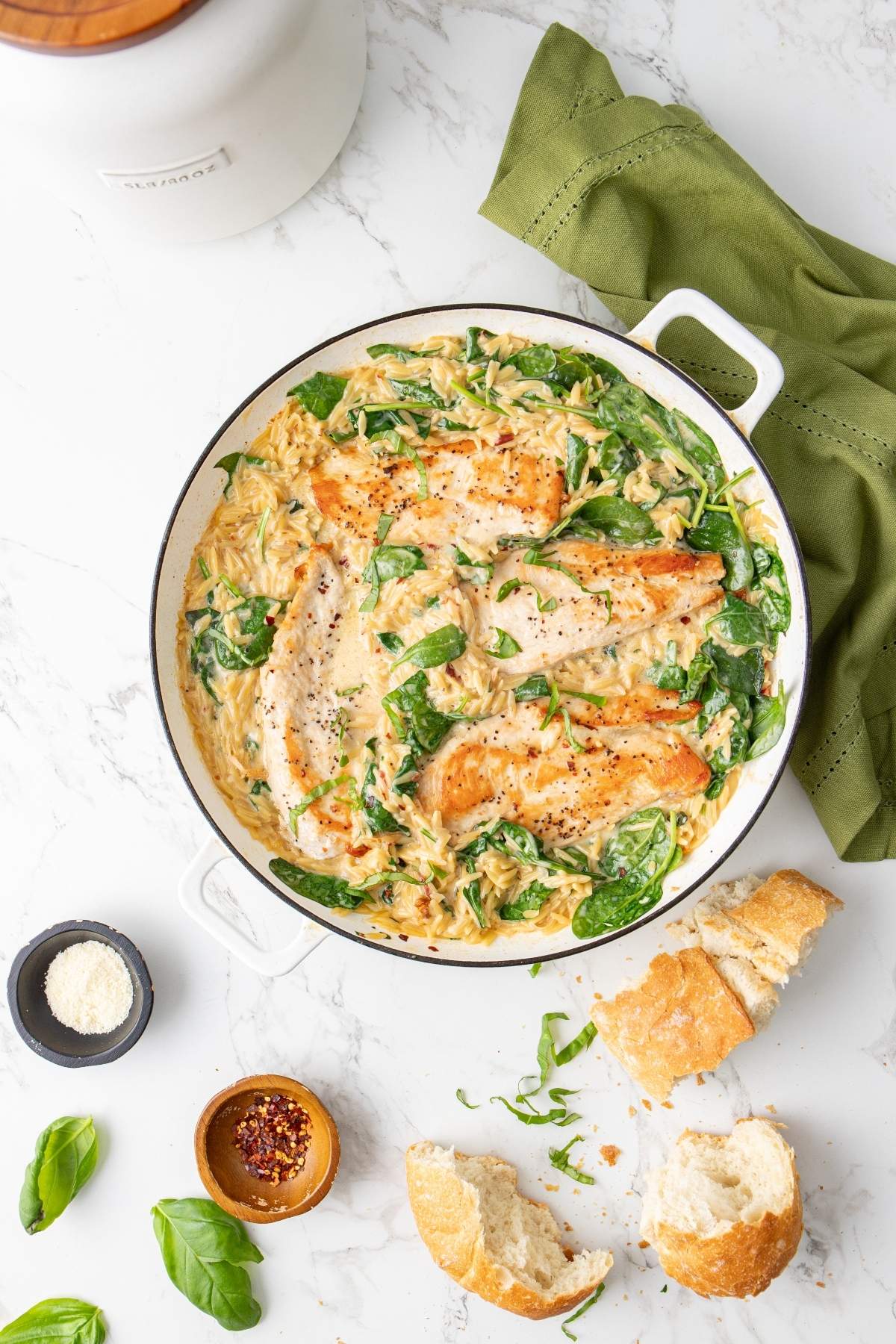 A white pot filled with creamy chicken and spinach orzo dish, garnished with black pepper. Fresh bread, red pepper flakes, and grated cheese are placed nearby on a white marble surface. A green cloth is seen on the side.