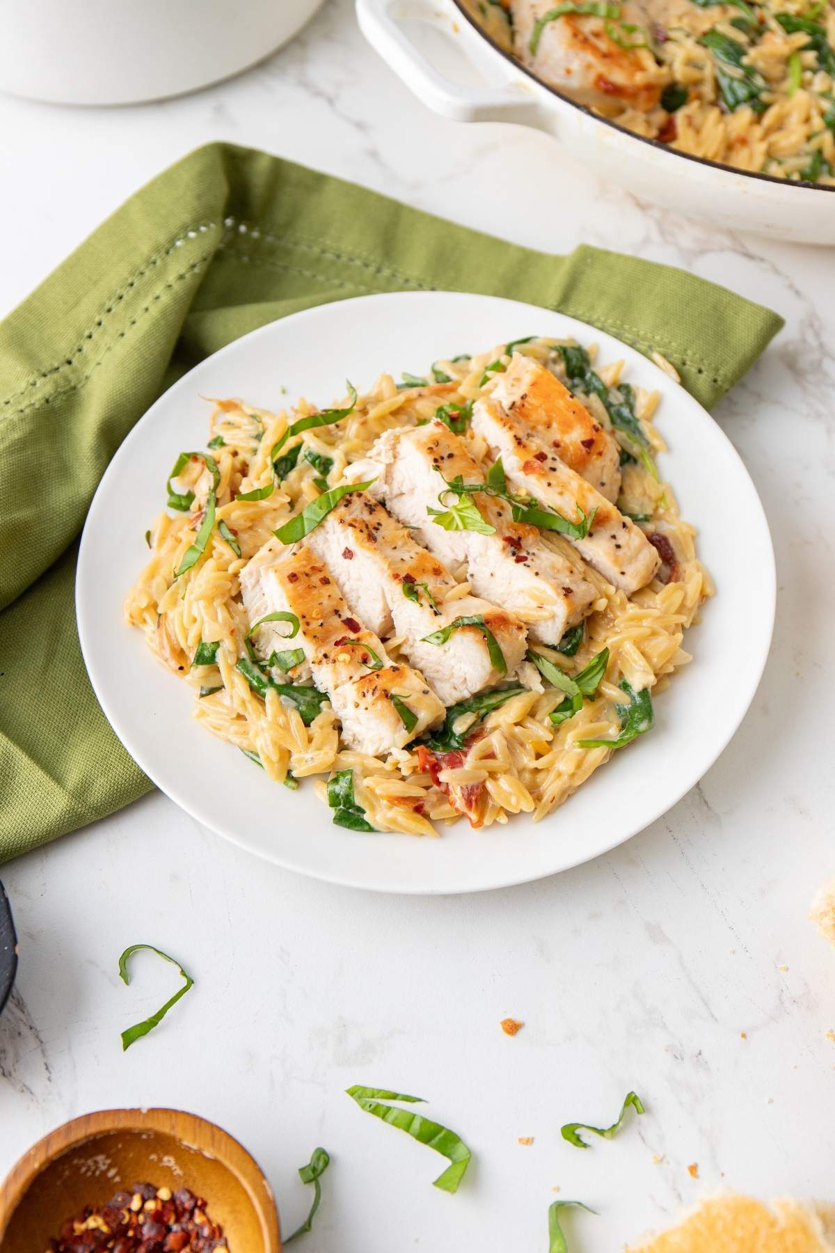 A dish of Italian chicken orzo with spinach and sliced grilled chicken breast, garnished with herbs, is served on a white plate next to a green napkin.